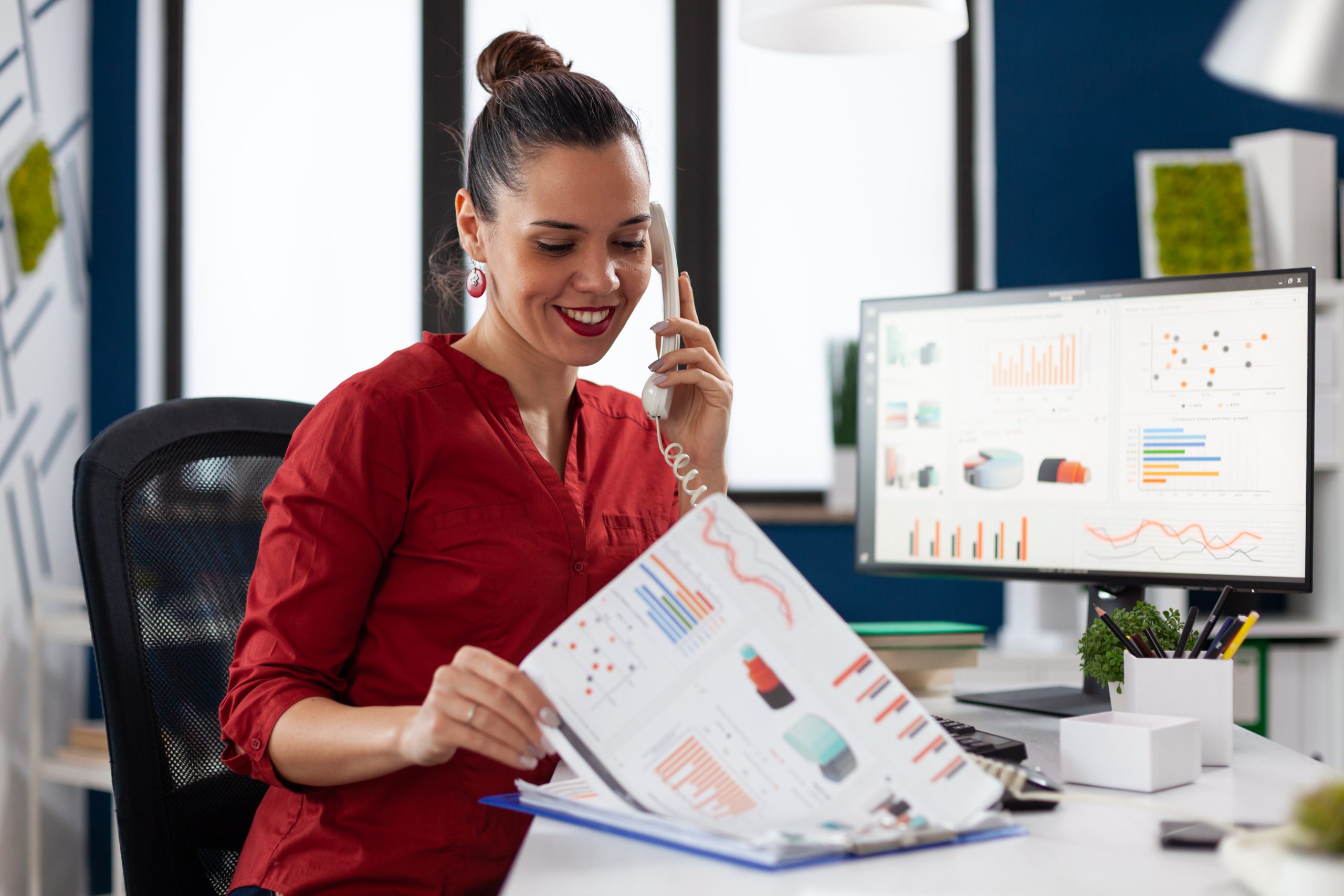 Eine Geschäftsfrau in roter Bluse sitzt in ihrem Büro und lächelt, während sie telefoniert und Finanzberichte mit bunten Diagrammen analysiert. Auf ihrem Schreibtisch stehen ein Monitor, auf dem Statistiken und visuelle Daten angezeigt werden, sowie organisierte Büromaterialien. Die moderne, professionelle Umgebung verfügt über ein raffiniertes Dekor und eine ausgewogene Beleuchtung.