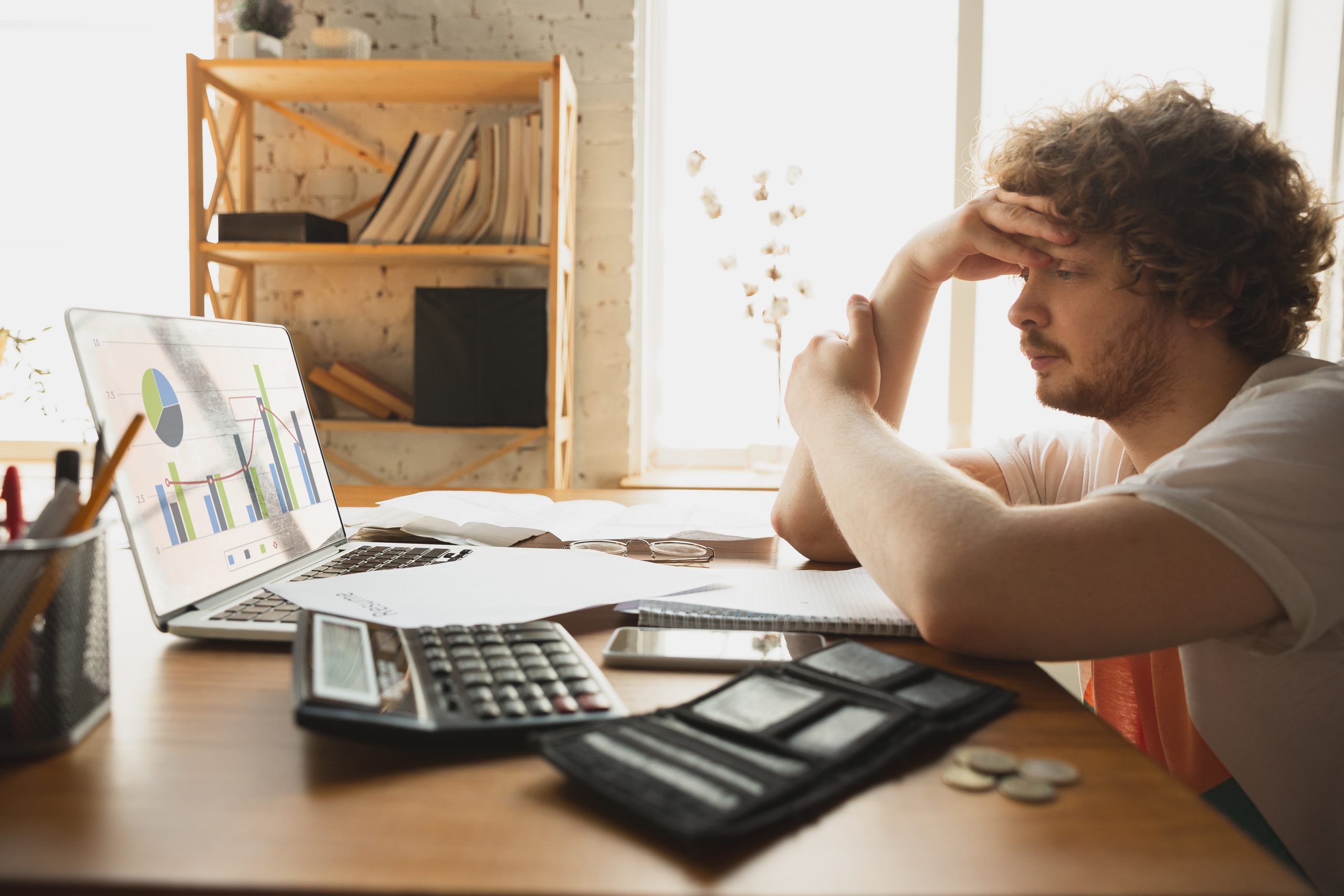 Ein müder, besorgt aussehender junger Mann sitzt an seinem Schreibtisch und stützt seinen Kopf auf seine Hand, während er auf einen Laptop mit Finanzdiagrammen starrt. Auf dem Schreibtisch liegen ein Taschenrechner, ein Notizblock, einige Münzen und ein offenes Portemonnaie, was auf eine Beschäftigung mit finanziellen Angelegenheiten hindeutet. Im Hintergrund stehen ein Bücherregal mit Büchern und Dokumenten sowie ein vom Tageslicht beleuchtetes Fenster. Übersetzt mit DeepL.com (kostenlose Version)