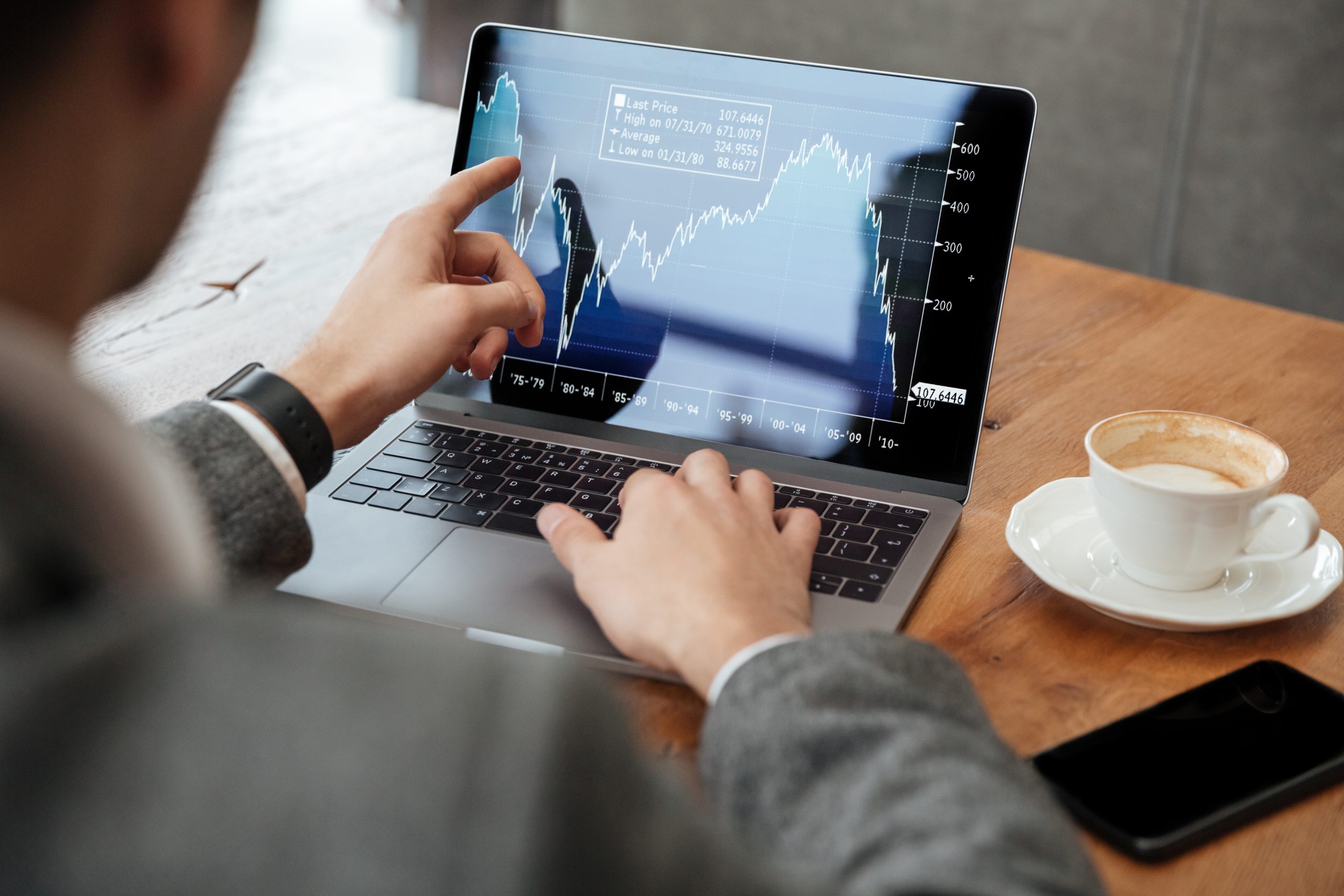 Ein Geschäftsmann in einem eleganten Anzug sitzt in einem Café und analysiert Finanzdiagramme auf seinem Laptop. Er zeigt auf den Bildschirm, auf dem ein Aktienchart mit den Schwankungen des Marktes angezeigt wird. Neben dem Computer stehen eine Tasse Kaffee und ein Smartphone auf dem Holztisch, was eine professionelle und anspruchsvolle Atmosphäre schafft.