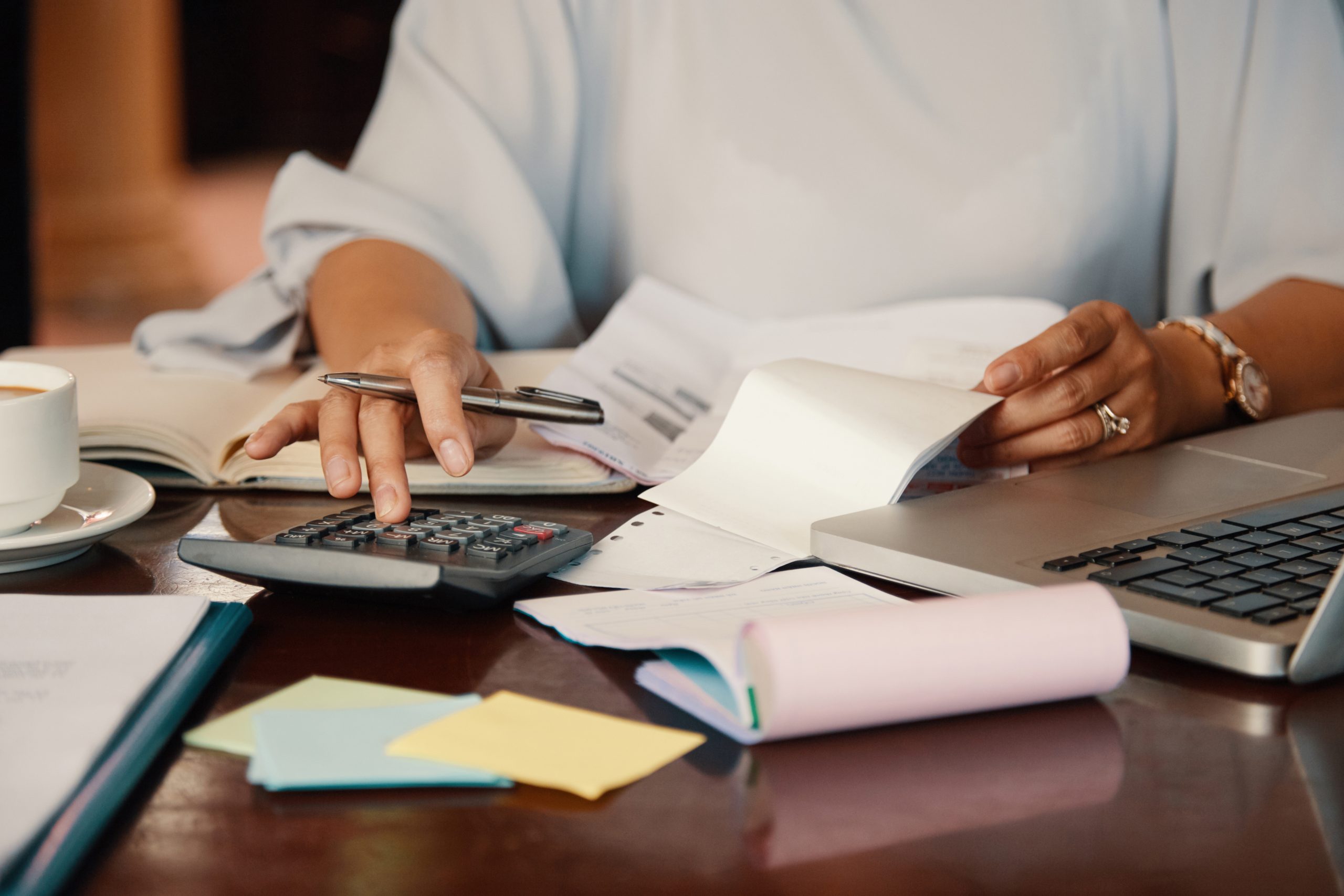Frau analysiert Konten und Finanzdokumente, während sie einen Taschenrechner und einen Laptop benutzt. Sie hält einen Stift und einen Notizblock in der Hand und konzentriert sich auf ihre Berechnungen. Der Schreibtisch ist mit farbigen Post-its, einer Tasse Kaffee und verstreuten Papieren ausgestattet, was auf ein Arbeitsumfeld schließen lässt, das auf Finanzplanung oder Buchhaltung ausgerichtet ist.