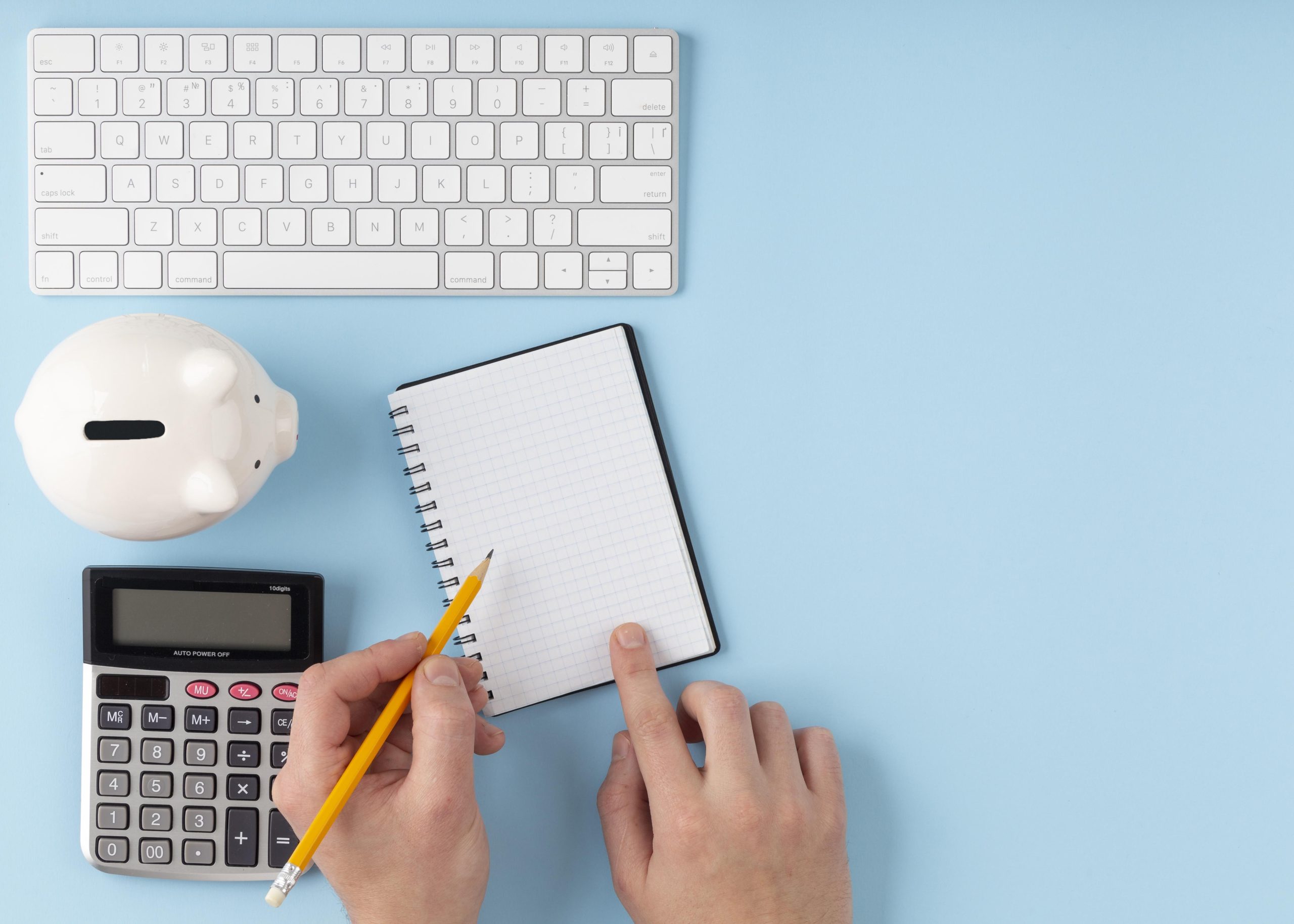 Ein Bild von einem organisierten Arbeitsbereich mit finanziellen Elementen auf blauem Hintergrund. In der Szene gibt es eine weiße Tastatur, ein Sparschwein, einen Taschenrechner, ein Notizbuch mit karierten Blättern und eine Hand, die einen Bleistift hält, bereit, Notizen zu machen. Die Komposition suggeriert Finanzplanung.