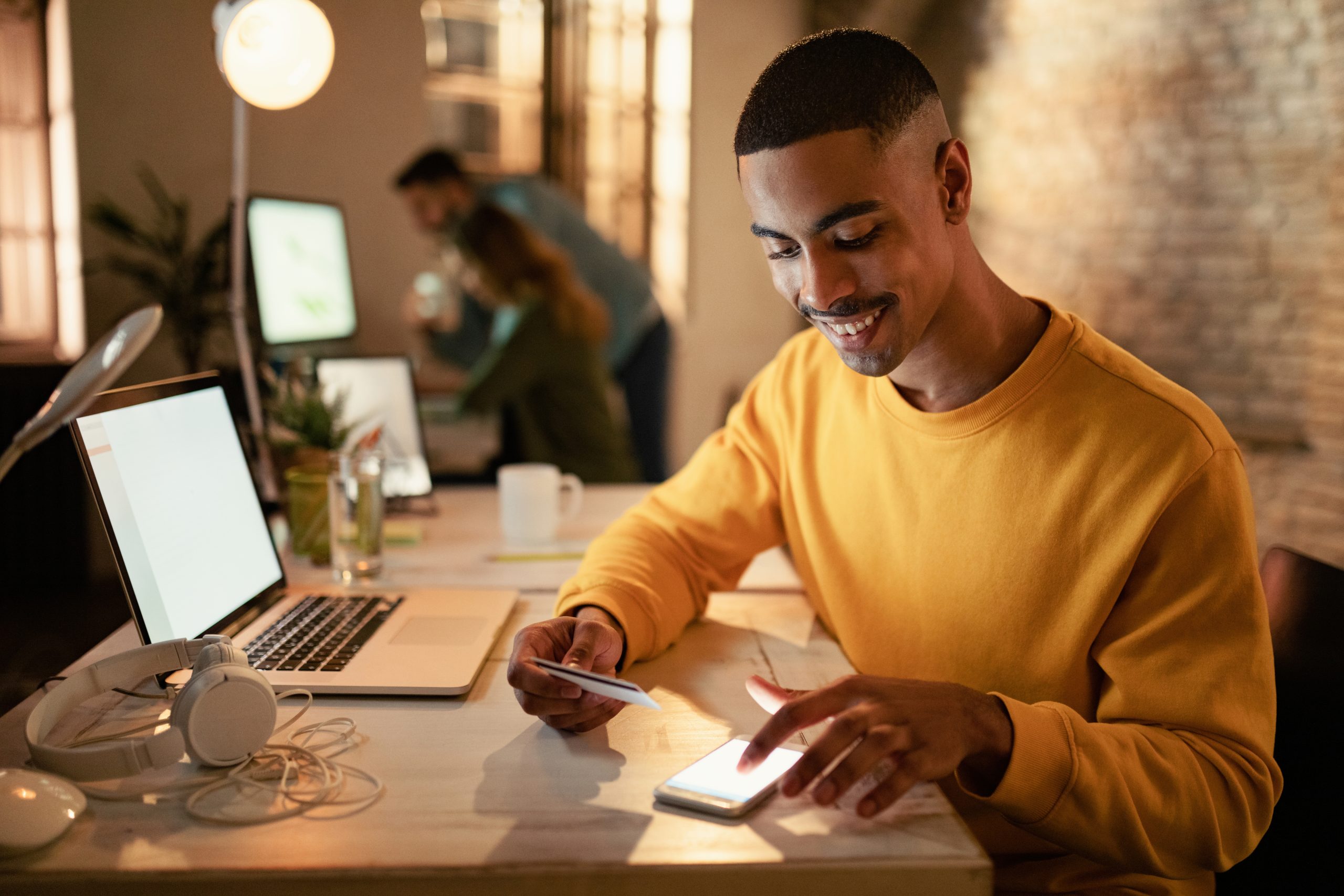Ein junger, lächelnder Mann in einem gelben Sweatshirt sitzt an einem Schreibtisch in einem modernen, gemütlichen Büro. Er hält eine Kreditkarte in der Hand, während er mit seinem Smartphone interagiert, als würde er eine Zahlung oder eine Online-Transaktion durchführen. Der Raum ist sanft beleuchtet, neben ihm stehen ein aufgeklappter Laptop, Kopfhörer und eine Tasse. Im Hintergrund arbeiten zwei Personen an einem anderen Tisch und schaffen so eine produktive und kollaborative Atmosphäre.