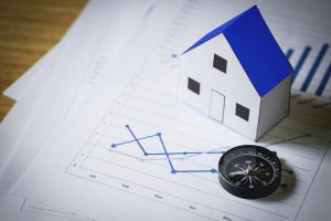 Das Bild zeigt ein Modell eines Miniaturhauses mit blauem Dach, das über Finanzdiagrammen und Dokumenten mit Daten und Statistiken steht. Neben dem Haus steht ein Kompass, der Orientierung und strategische Entscheidungen symbolisiert. Die Szene steht für Konzepte wie den Immobilienmarkt, Immobilieninvestitionen und die Analyse von Wirtschaftstrends.