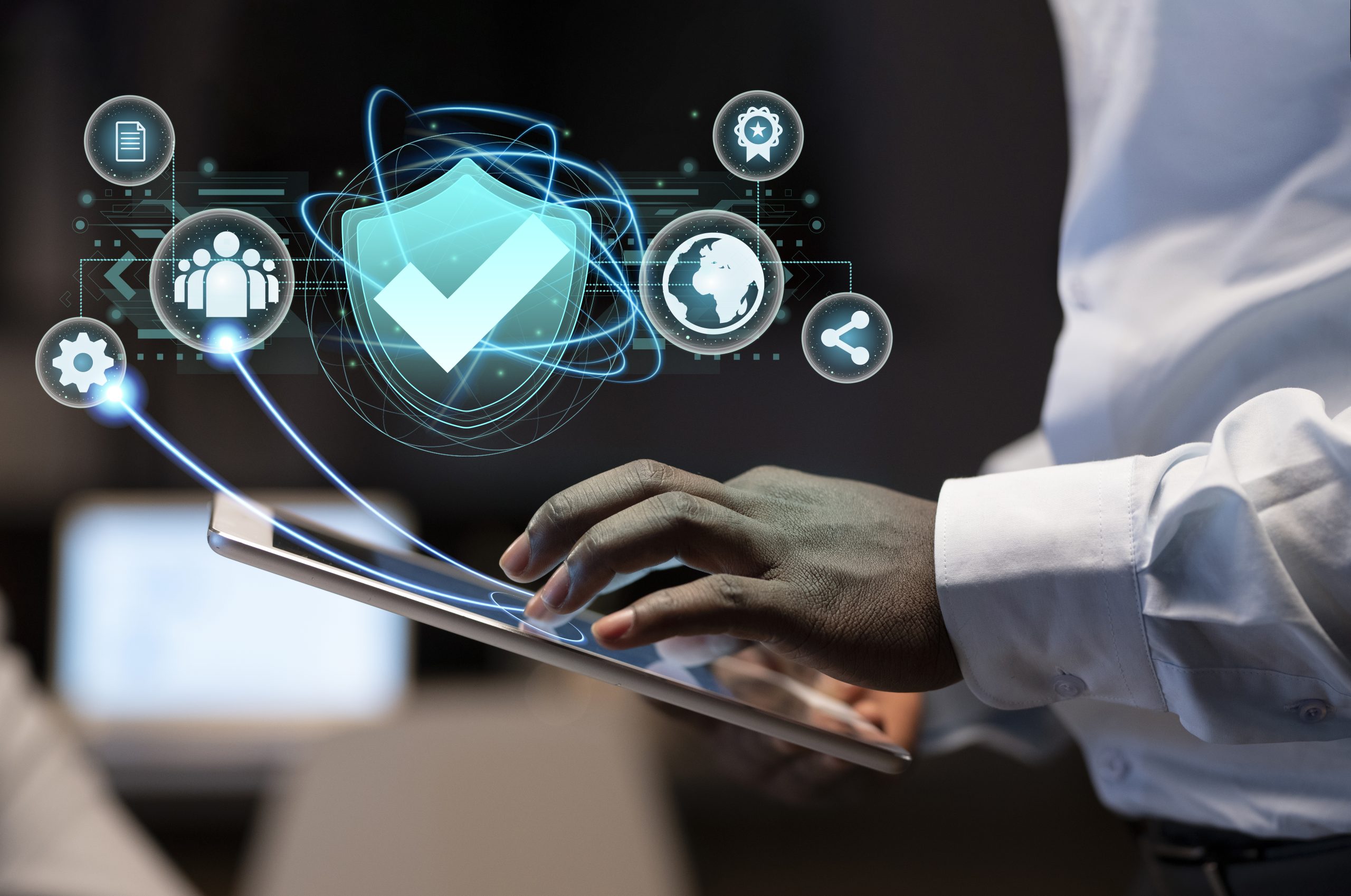 Ein Geschäftsmann demonstriert die Bedeutung der Cybersicherheit auf seinem Tablet, das ein Schutzschild mit verschiedenen Sicherheitssymbolen zeigt, die den Schutz von Daten und Systemen symbolisieren. Der Hintergrund mit einem modernen Büro deutet auf die Anwendung von Sicherheit in Unternehmensumgebungen hin.