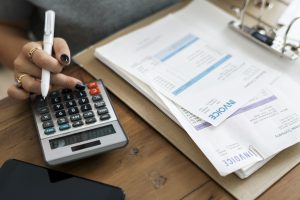 Eine Frau führt mit einem Taschenrechner finanzielle Berechnungen durch und hält dabei einen Stift in der Hand. Auf dem Holztisch liegen mehrere Rechnungen und Dokumente in einem Aktenordner, was auf eine Tätigkeit in der Buchhaltung oder Finanzverwaltung schließen lässt. Daneben liegt ein Smartphone, das die professionelle Atmosphäre vervollständigt.
