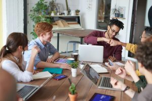 Eine Gruppe junger Menschen arbeitet gemeinsam an einem Holztisch in einem modernen Büro; zwei Männer geben sich einen Fistbump, während andere lachen, diskutieren und Laptops, Notizbücher und Tablets benutzen – die Szene vermittelt Teamarbeit, Kreativität und ein positives Arbeitsumfeld.