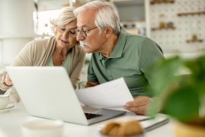 Ein älteres Paar sitzt gemeinsam am Tisch und schaut konzentriert auf einen Laptop, während der Mann ein Dokument in der Hand hält; sie besprechen vermutlich ihre Finanzen oder Altersvorsorge in einem hellen, gemütlichen Zuhause.