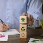Ein Mann im blauen Hemd stapelt Holzwürfel mit Symbolen für Recycling, Kreislaufwirtschaft und Umweltschutz auf einem Schreibtisch – Konzept nachhaltiger Entwicklung und ökologischer Verantwortung.