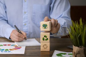 Ein Mann im blauen Hemd stapelt Holzwürfel mit Symbolen für Recycling, Kreislaufwirtschaft und Umweltschutz auf einem Schreibtisch – Konzept nachhaltiger Entwicklung und ökologischer Verantwortung.
