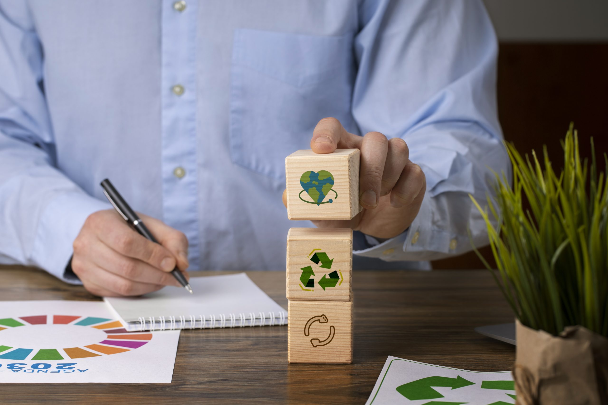 Ein Mann im blauen Hemd stapelt Holzwürfel mit Symbolen für Recycling, Kreislaufwirtschaft und Umweltschutz auf einem Schreibtisch – Konzept nachhaltiger Entwicklung und ökologischer Verantwortung.