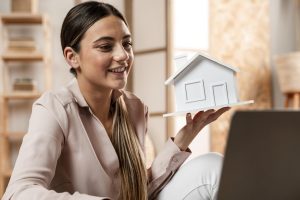 Eine lächelnde Frau hält ein weißes Modellhaus in der Hand und schaut auf einen Laptop – symbolisch für Immobilienberatung, Hauskauf oder Online-Immobiliengeschäfte.