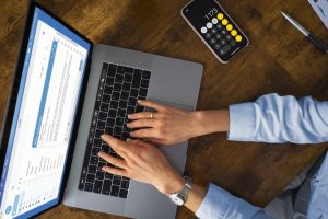 Eine Person tippt auf einem Laptop, während auf dem Bildschirm eine E-Mail-Anwendung geöffnet ist und daneben ein Smartphone mit geöffneter Taschenrechner-App liegt. Die Szene verdeutlicht den Einfluss der Digitalisierung auf die Steuererklärung, da moderne Technologien zunehmend eine effiziente und papierlose Abwicklung steuerlicher Angelegenheiten ermöglichen.