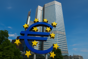 Vor dem modernen Hochhaus des Europäischen Zentralbanksitzes in Frankfurt steht das große blaue Euro-Symbol, umgeben von gelben Sternen der Europäischen Union. Die Szene symbolisiert die wirtschaftliche Stabilität und Herausforderungen wie die Inflation in der Eurozone, die durch geldpolitische Maßnahmen der EZB beeinflusst werden.