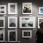 Eine Frau betrachtet aufmerksam eine Wand voller gerahmter Fotografien und Kunstwerke in einer Galerie. Die Szene symbolisiert das wachsende Interesse am Investieren in Kunst und Sammlerstücke in Deutschland, da solche Ausstellungen potenziellen Investoren und Kunstliebhabern eine Vielzahl an Möglichkeiten bieten, einzigartige Werke zu entdecken und in deren Wertentwicklung zu investieren.