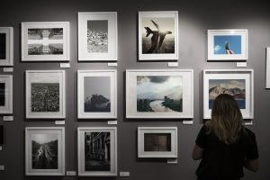 Eine Frau betrachtet aufmerksam eine Wand voller gerahmter Fotografien und Kunstwerke in einer Galerie. Die Szene symbolisiert das wachsende Interesse am Investieren in Kunst und Sammlerstücke in Deutschland, da solche Ausstellungen potenziellen Investoren und Kunstliebhabern eine Vielzahl an Möglichkeiten bieten, einzigartige Werke zu entdecken und in deren Wertentwicklung zu investieren.