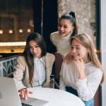 Drei junge Frauen sitzen gemeinsam an einem Tisch in einem modernen Café und schauen lächelnd auf einen Laptop-Bildschirm. Die Szene symbolisiert den Austausch und die Zusammenarbeit von Kleinunternehmern, die digitale Technologien nutzen, um ihre Geschäftsstrategien zu entwickeln und ihre unternehmerischen Ziele erfolgreich zu verfolgen.