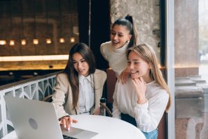 Drei junge Frauen sitzen gemeinsam an einem Tisch in einem modernen Café und schauen lächelnd auf einen Laptop-Bildschirm. Die Szene symbolisiert den Austausch und die Zusammenarbeit von Kleinunternehmern, die digitale Technologien nutzen, um ihre Geschäftsstrategien zu entwickeln und ihre unternehmerischen Ziele erfolgreich zu verfolgen.