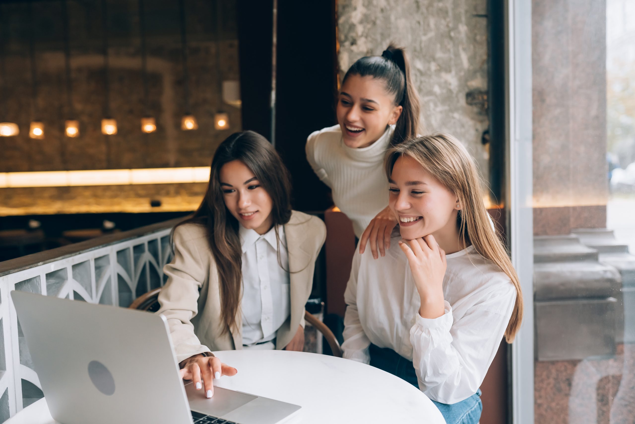 Drei junge Frauen sitzen gemeinsam an einem Tisch in einem modernen Café und schauen lächelnd auf einen Laptop-Bildschirm. Die Szene symbolisiert den Austausch und die Zusammenarbeit von Kleinunternehmern, die digitale Technologien nutzen, um ihre Geschäftsstrategien zu entwickeln und ihre unternehmerischen Ziele erfolgreich zu verfolgen.