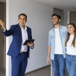 Ein Immobilienmakler zeigt einem jungen Paar eine moderne Wohnung, während er freundlich auf den Raum deutet und ein Tablet in der Hand hält. Diese Szene veranschaulicht den Prozess der Besichtigung von Mietimmobilien in Deutschland, bei dem potenzielle Mieter gemeinsam mit Maklern passende Objekte auswählen und wichtige Informationen zur Anmietung erhalten.