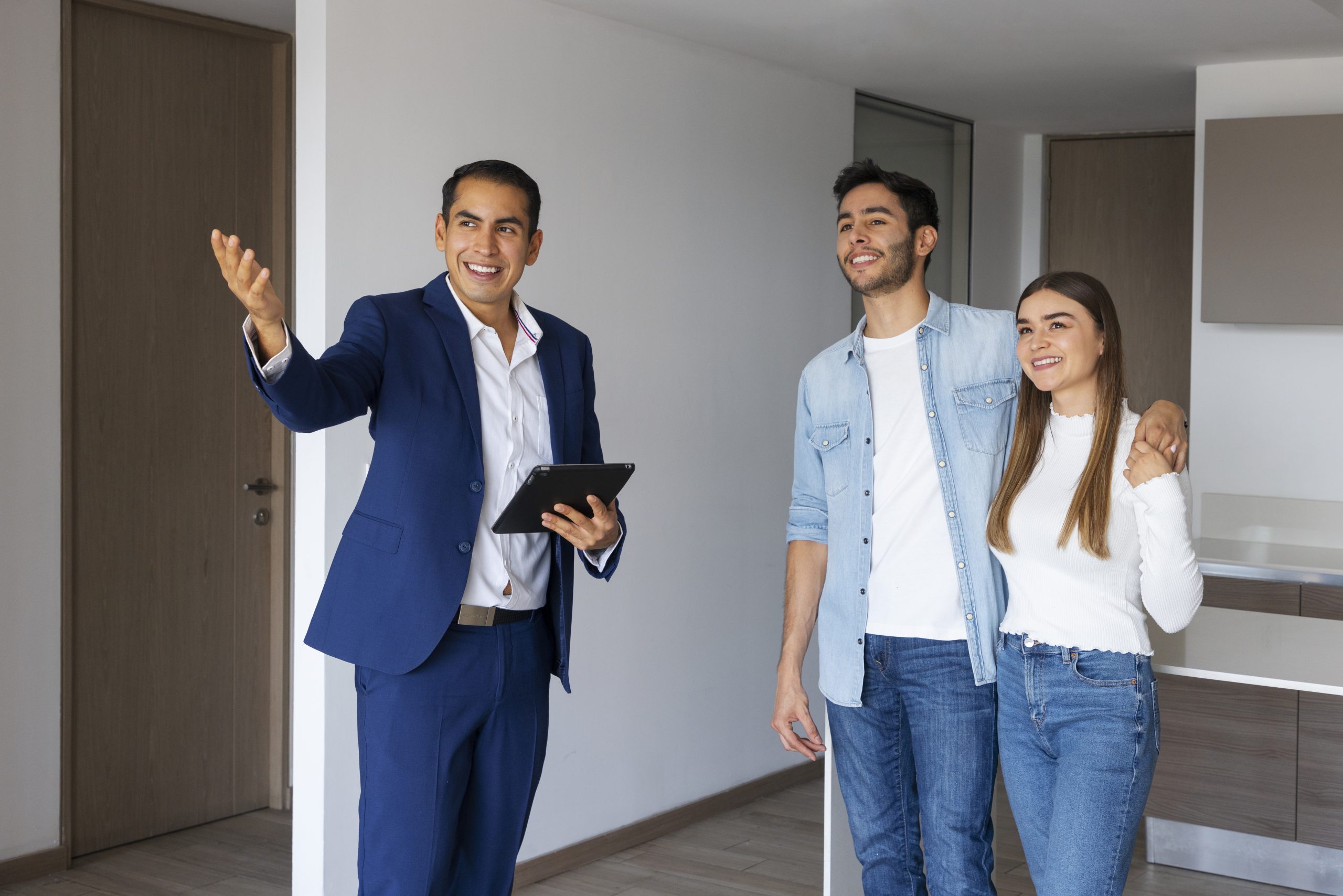 Ein Immobilienmakler zeigt einem jungen Paar eine moderne Wohnung, während er freundlich auf den Raum deutet und ein Tablet in der Hand hält. Diese Szene veranschaulicht den Prozess der Besichtigung von Mietimmobilien in Deutschland, bei dem potenzielle Mieter gemeinsam mit Maklern passende Objekte auswählen und wichtige Informationen zur Anmietung erhalten.