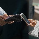 Eine Person hält ein Smartphone an ein kontaktloses Kartenlesegerät, um eine Zahlung durchzuführen. Diese Szene illustriert den Einsatz moderner Zahlungsmittel, die dank digitaler Technologien schnelle und sichere Transaktionen ermöglichen und zunehmend das traditionelle Bargeld im Alltag ersetzen.