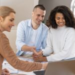 Ein lächelndes Paar sitzt gemeinsam mit einer Beraterin an einem Tisch, auf dem Vertragsunterlagen und ein Laptop liegen. Alle drei schauen auf den Bildschirm und diskutieren offenbar wichtige Details. Die Szene symbolisiert die persönliche Beratung und vertrauensvolle Zusammenarbeit, die typisch für genossenschaftlichen Banken ist und den Mitgliedern individuelle Lösungen bietet.