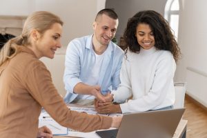 Ein lächelndes Paar sitzt gemeinsam mit einer Beraterin an einem Tisch, auf dem Vertragsunterlagen und ein Laptop liegen. Alle drei schauen auf den Bildschirm und diskutieren offenbar wichtige Details. Die Szene symbolisiert die persönliche Beratung und vertrauensvolle Zusammenarbeit, die typisch für genossenschaftlichen Banken ist und den Mitgliedern individuelle Lösungen bietet.