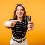 Fröhliche junge Frau vor gelbem Hintergrund zeigt stolz auf eine schwarze Kreditkarte – symbolisches Bild für moderne Finanzvorteile wie Cashback bei Kartenzahlungen.