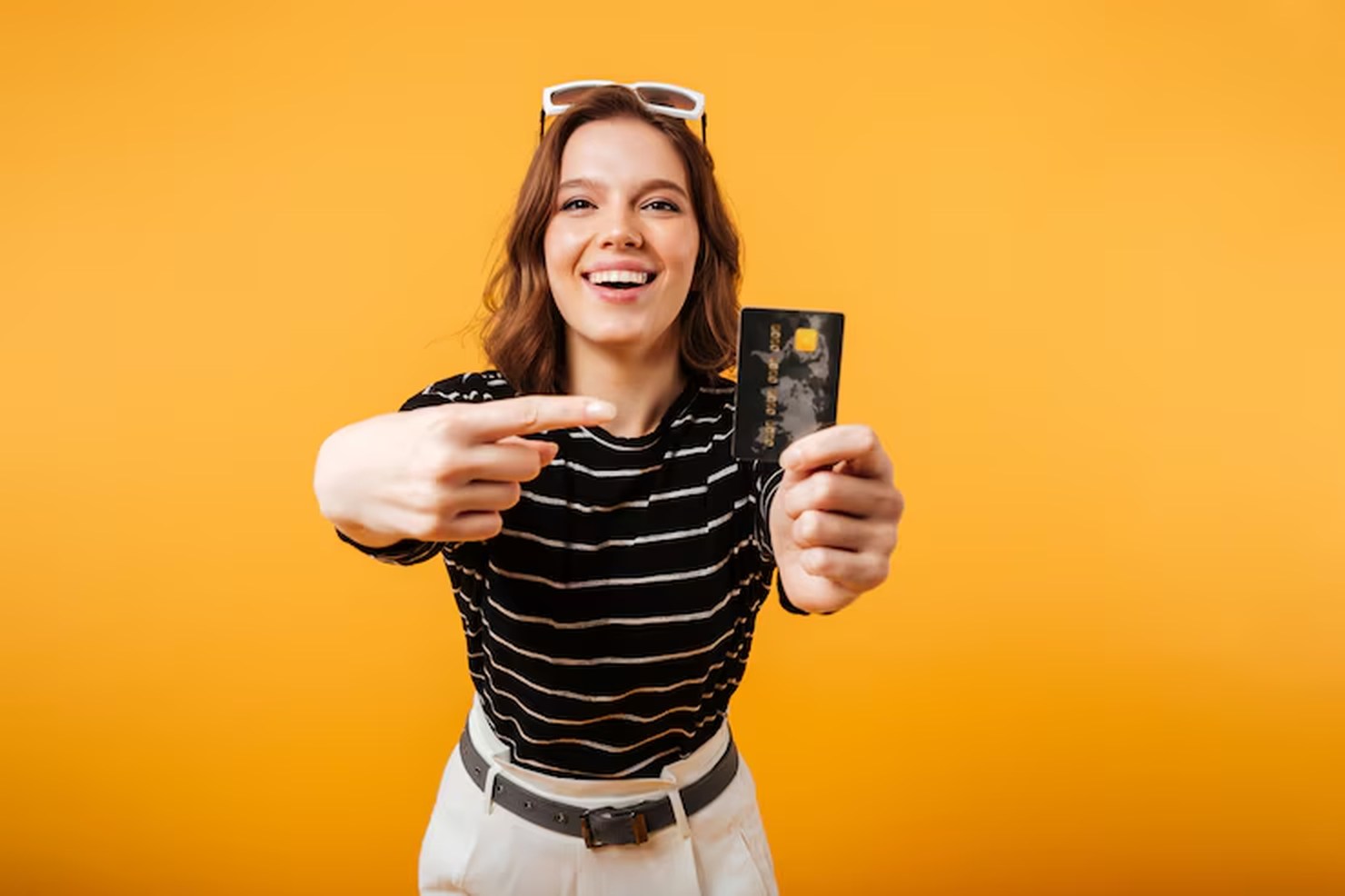 Fröhliche junge Frau vor gelbem Hintergrund zeigt stolz auf eine schwarze Kreditkarte – symbolisches Bild für moderne Finanzvorteile wie Cashback bei Kartenzahlungen.