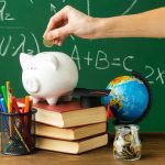 Symbolbild für finanzielle Bildung: Hand wirft Münze in ein Sparschwein, umgeben von Schulbüchern, Schreibmaterial, Globus und Tafel mit mathematischen Formeln im Hintergrund.