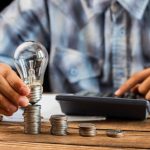 Ein Mann stapelt Münzen auf einem Holztisch und hält dabei eine Glühbirne, während er mit der anderen Hand einen Taschenrechner bedient – symbolische Darstellung für den sorgfältigen Umgang mit Finanzen in Zeiten der Energiekrise.
