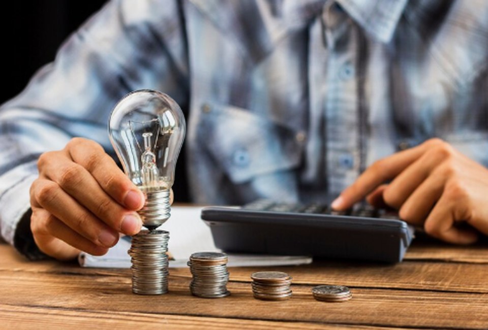 Ein Mann stapelt Münzen auf einem Holztisch und hält dabei eine Glühbirne, während er mit der anderen Hand einen Taschenrechner bedient – symbolische Darstellung für den sorgfältigen Umgang mit Finanzen in Zeiten der Energiekrise.