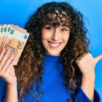 Eine junge Frau mit lockigem Haar lächelt fröhlich, während sie mehrere Euro-Banknoten in der einen Hand hält und mit der anderen Hand stolz nach hinten zeigt – Symbol für finanziellen Erfolg und Selbstvertrauen einer neuen Generation.