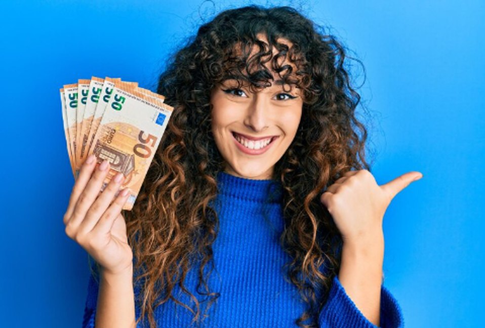 Eine junge Frau mit lockigem Haar lächelt fröhlich, während sie mehrere Euro-Banknoten in der einen Hand hält und mit der anderen Hand stolz nach hinten zeigt – Symbol für finanziellen Erfolg und Selbstvertrauen einer neuen Generation.