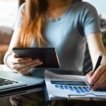Eine Frau arbeitet an einem Schreibtisch mit einem Laptop, einem Tablet und einem Finanzbericht mit Diagrammen. Sie macht sich Notizen, während sie Daten analysiert – ein typisches Szenario im Bereich persönliche Finanzen.