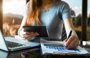 Eine Frau arbeitet an einem Schreibtisch mit einem Laptop, einem Tablet und einem Finanzbericht mit Diagrammen. Sie macht sich Notizen, während sie Daten analysiert – ein typisches Szenario im Bereich persönliche Finanzen.