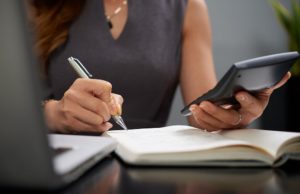 Frau berechnet Finanzen mit Notizbuch und Taschenrechner am Schreibtisch, Symbol für Planung und Verwaltung der Sozialversicherung.