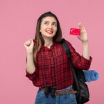Junge Frau mit Rucksack und Fernglas hält freudig eine rote Bankkarte in der Hand, als Symbol für finanzielle Freiheit und die Vorteile von Karten für Studierende.