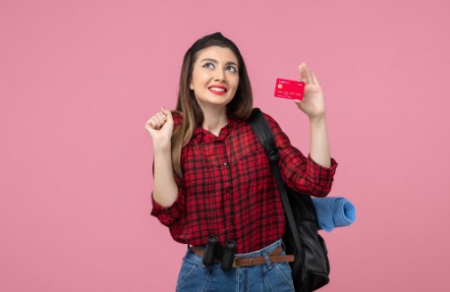 Junge Frau mit Rucksack und Fernglas hält freudig eine rote Bankkarte in der Hand, als Symbol für finanzielle Freiheit und die Vorteile von Karten für Studierende.