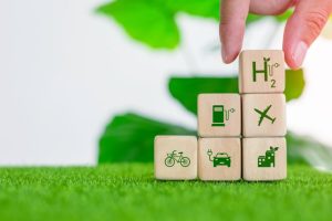 Holzwürfel mit Symbolen für Fahrrad, Elektroauto, Wasserstoff, Flugzeug und nachhaltige Gebäude auf grüner Wiese – eine Darstellung von Innovation und Nachhaltigkeit in der Mobilität und Energieversorgung.