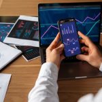 Person analysiert Finanzdiagramme auf Laptop und Smartphone, um Investitionsstrategien mit Hilfe von digitalen Werkzeugen besser zu planen und zu überwachen.