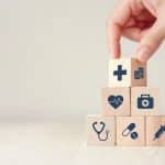 Hand, die Holzblöcke mit Symbolen für Gesundheit und Finanzen zu einer Pyramide stapelt, was Stabilität, Sicherheit und die Bedeutung einer vorausschauenden Finanzplanung (Finanzplanung) symbolisiert.