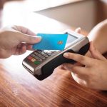 Person, die eine blaue Karte kontaktlos an ein Zahlungsterminal hält, symbolisiert moderne Zahlungsmethoden und den Einsatz von Karten mit Versicherungsleistungen für mehr Sicherheit und Komfort im Alltag.